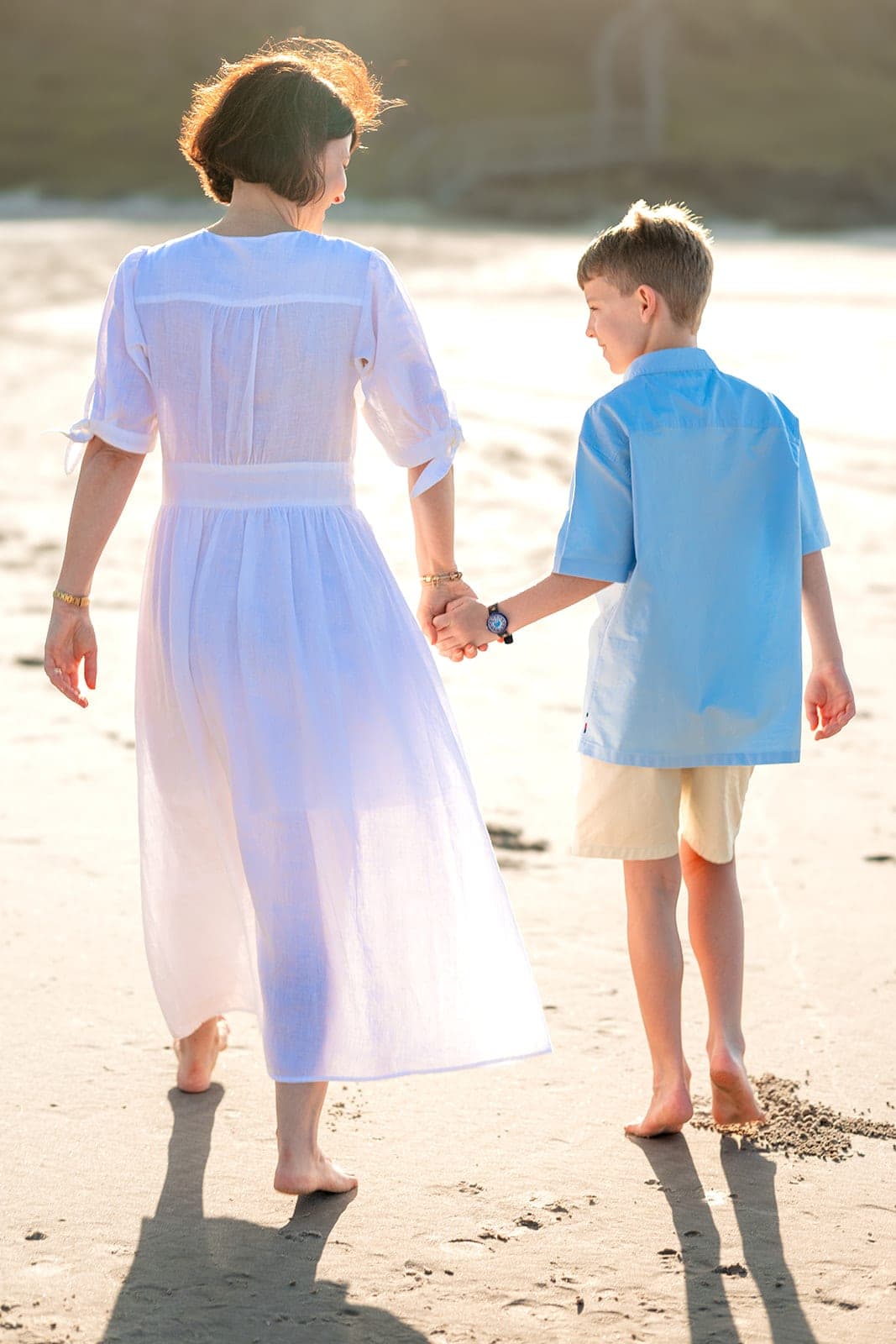 Mum in a white dress walking hand in hand with her son on the sand