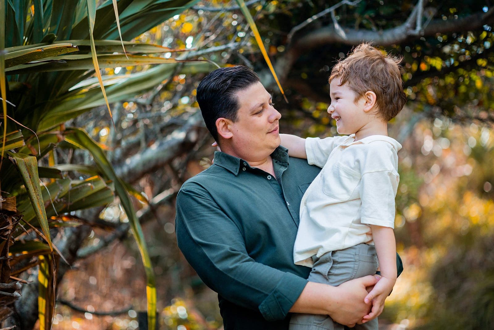Father holding his young son, both laughing among palm fronds