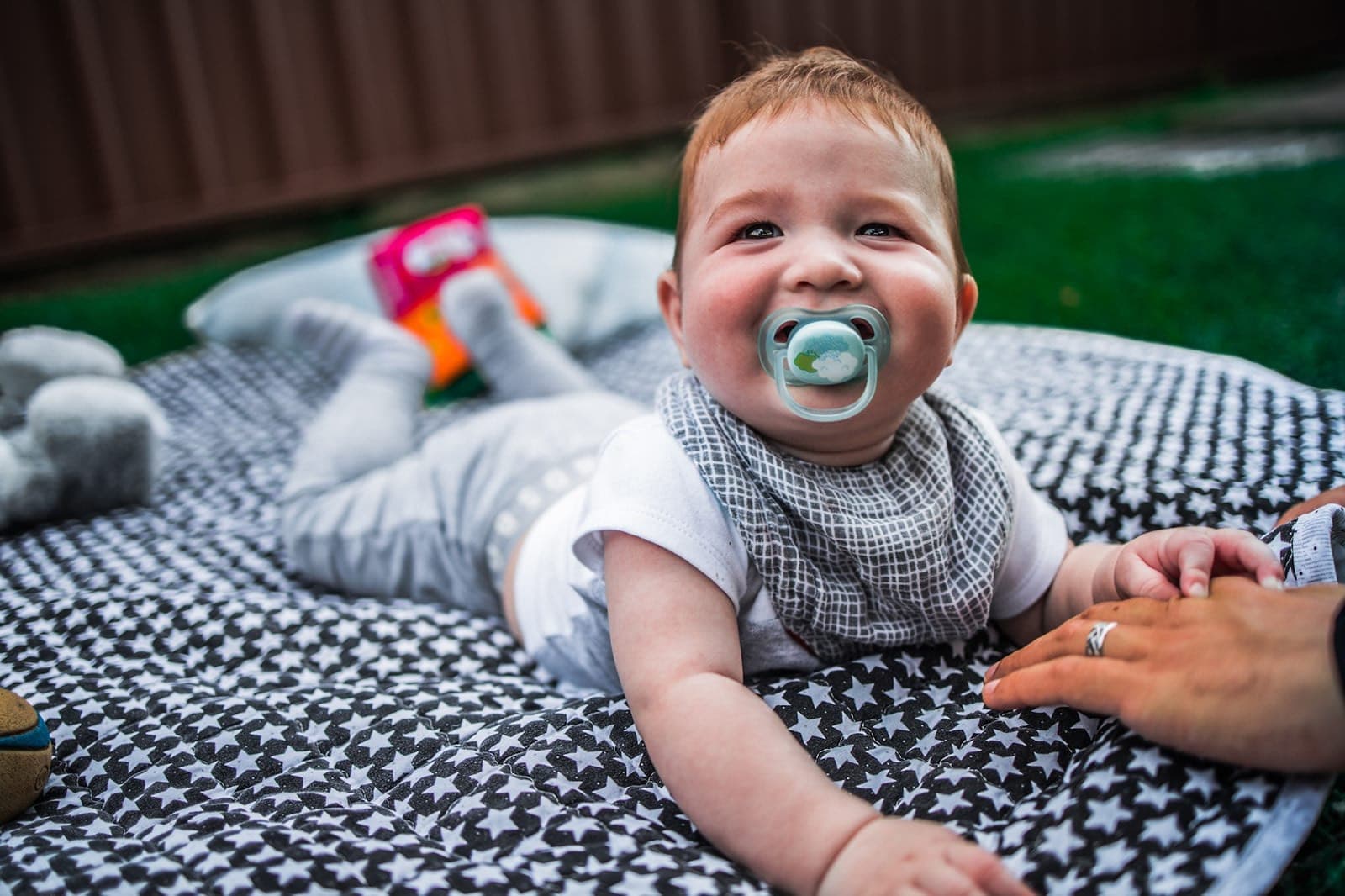 Smiling baby with a dummy lying on a picnic rug in the garden