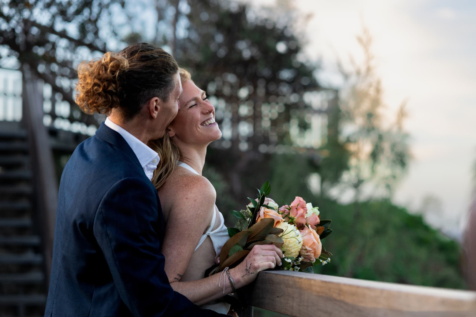 Bride and groom laughing together against the timber railing of a coastal lookout