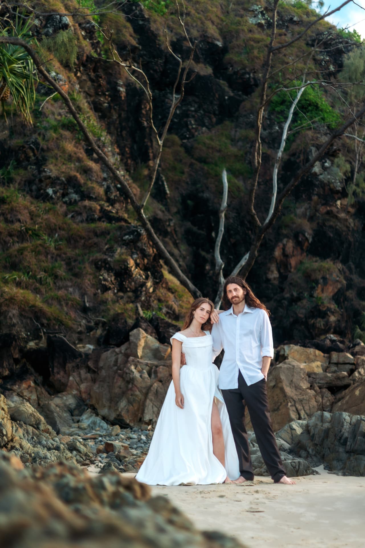 Bride and groom standing close beneath coastal cliffs and scrub