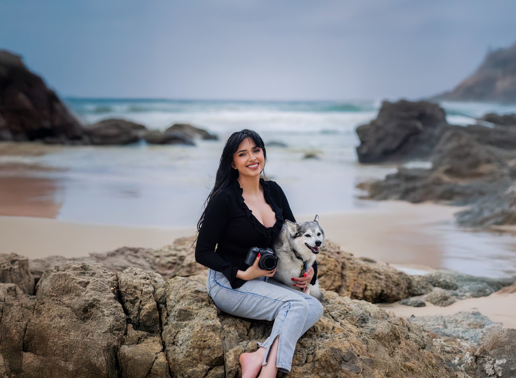 Heather Hilton sitting on beach rocks holding her camera beside her husky
