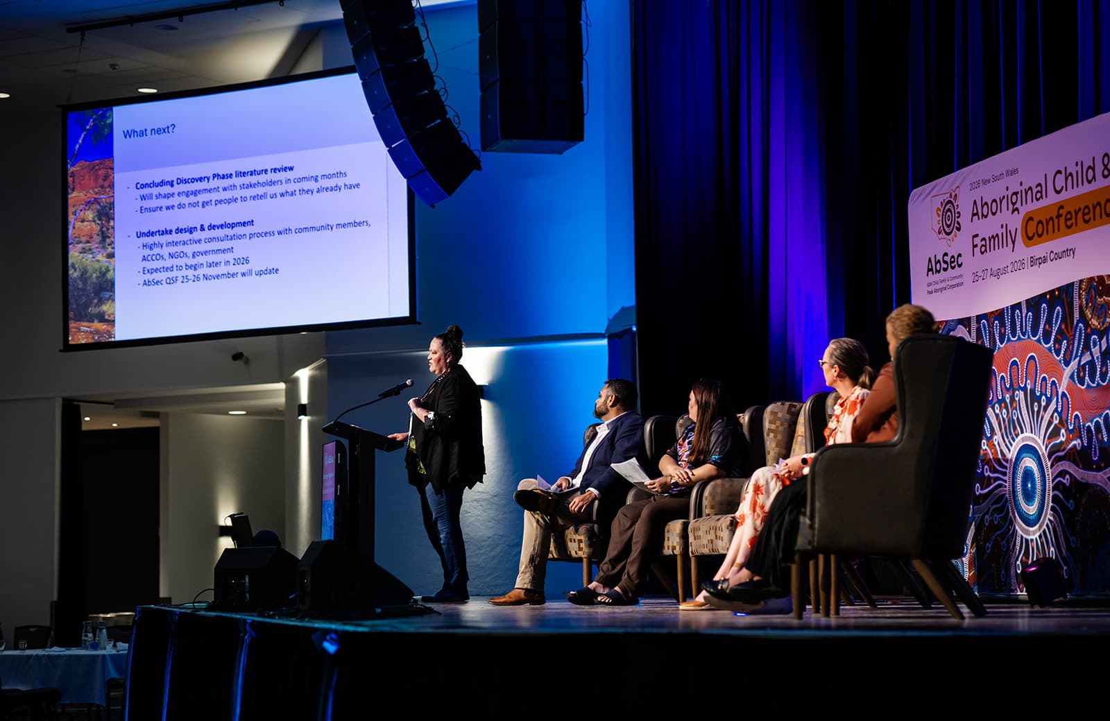 A speaker at the lectern beside a seated panel and the presentation screen