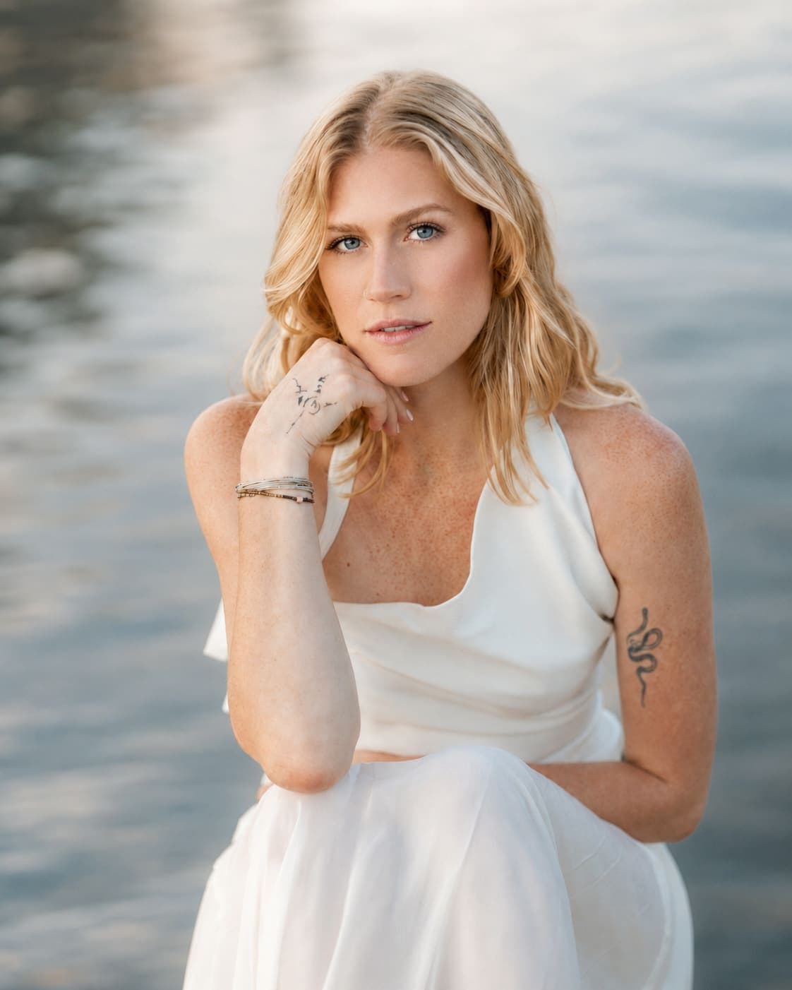 Bride with blonde hair seated at the water’s edge in soft light