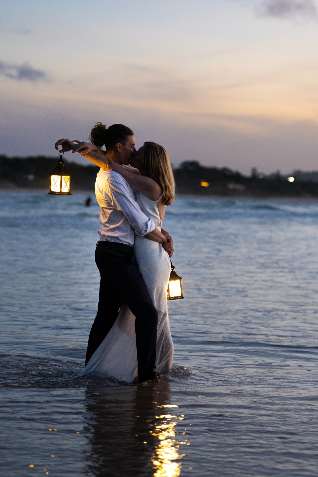 Couple embracing knee-deep in the ocean at dusk holding lanterns