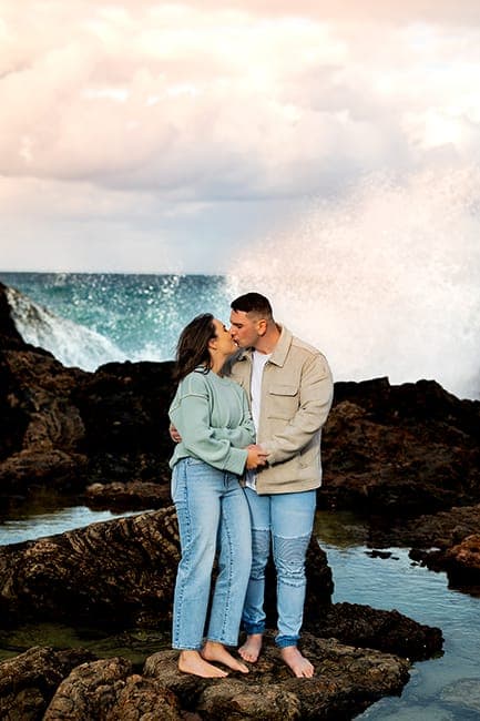 Couple embracing on the rocks as a wave bursts behind them