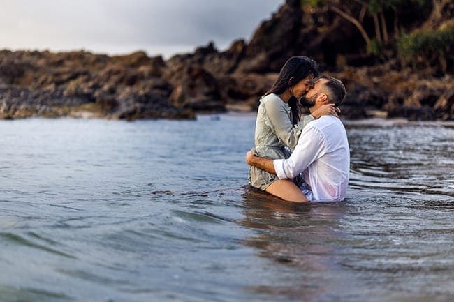 Couple embracing waist-deep in calm water below a rocky headland