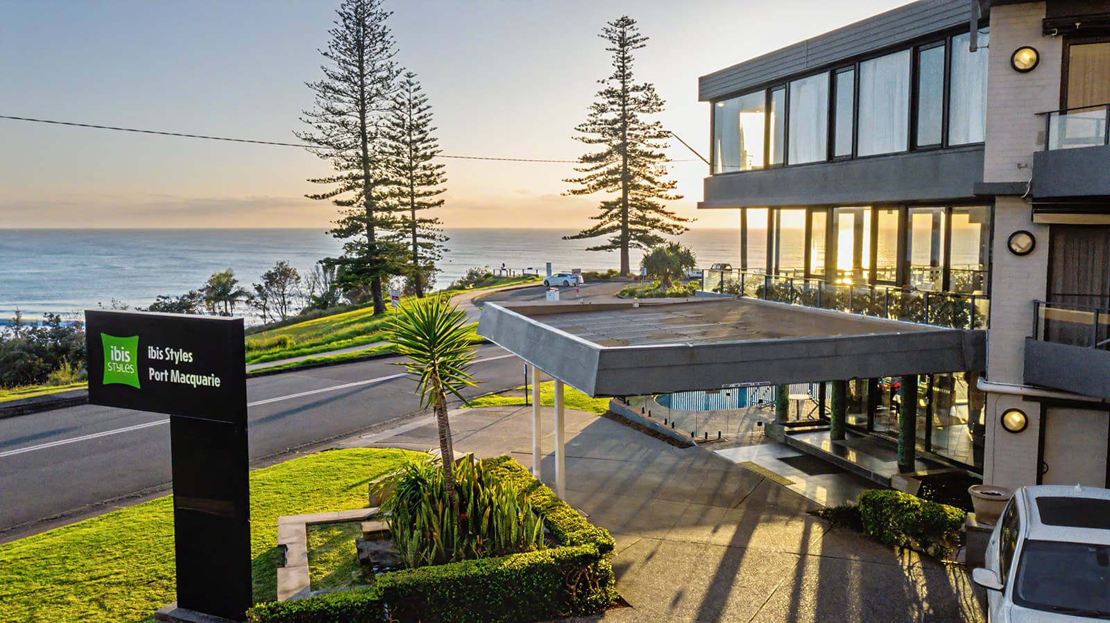 Ibis Styles Port Macquarie entrance and sign at sunrise with Norfolk pines and the ocean behind