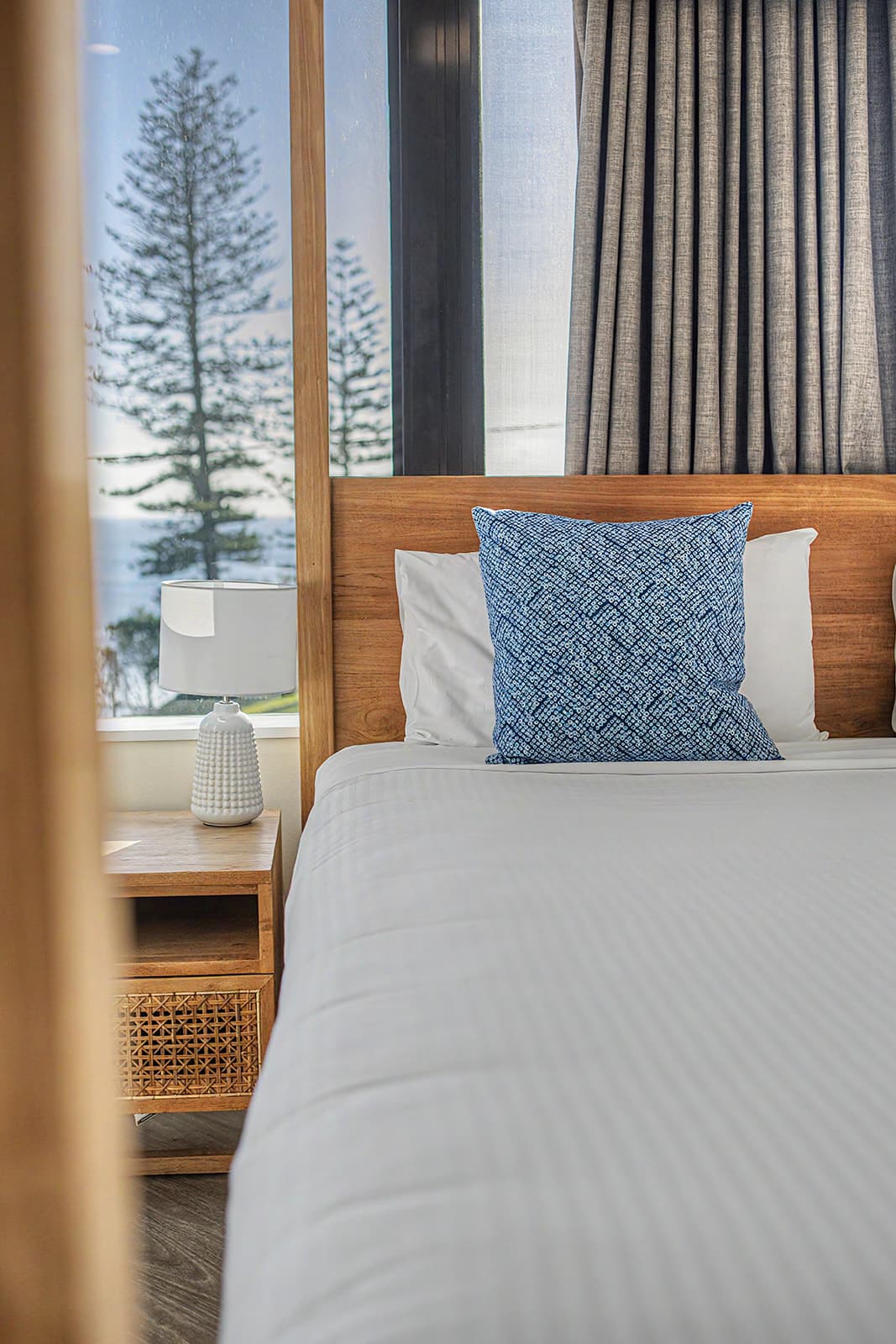 Bed and bedside lamp framed by a window looking out to a Norfolk pine and the sea